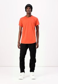 Oransje bomull t-skjorte med rulleterminger, kombinert med svarte bukser og hvite sneakers. Modell som bærer en svart lue med logo.