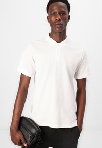 Polo shirt bianco in cotone con colletto classico, maniche corte e un piccolo logo sul petto. Tenuto in mano con una borsa nera.