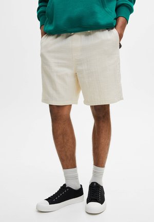 Hellbeige Shorts aus strukturiertem Stoff mit einem elastischen Bund und seitlichen Taschen, kombiniert mit schwarzen Sneakern und grauen Knöchelsocken.