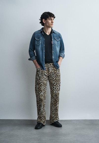 Νεαρός άνδρας που στέκεται με τα χέρια στις τσέπες, φορώντας τζιν πουκάμισο, μαύρο μπλουζάκι, παντελόνι με λεοπάρ μοτίβο και μαύρα παπούτσια μπροστά σε έναν απλό τοίχο.