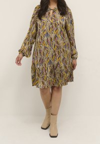 Robe à manches longues de couleur jaune moutarde ornée d'un motif de feuilles violet et bleu. Encolure en trou de serrure et jupe froncée, portée avec des bottines beiges.