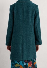 Cappotto di lana a spina di pesce color turchese indossato da una persona, con vista posteriore, mentre un gonna a fantasia astratta e colorata è visibile sotto l'orlo del cappotto.