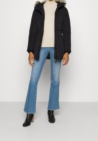 Person som bär en svart huvad parkas med pälsdetaljer, beige polotröja, ljusblå utsvängda jeans och svarta ankelboots med klack.