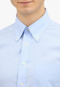 Jasnoniebieska koszula z guzikami, z kołnierzykiem zapinanym na guziki i kieszenią na piersi, noszona przez osobę z neutralnym wyrazem twarzy.