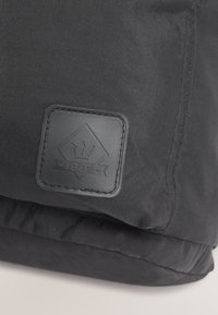 Tissu noir avec une pièce en cuir cousue représentant une forme de diamant, un symbole de trident, et le texte "1982-" embossé.