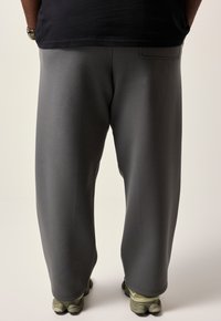 Sækkede grå sweatpants med en glat tekstur og elastisk talje. Har en baglomme og sidder over anklen, kombineret med olivengrønne sneakers.