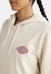 Kremowy hoodie z kieszenią z przodu. Posiada haftowany wzór oka w czerwonym, niebieskim i słonecznym motywie. Sznurki przy kapturze.