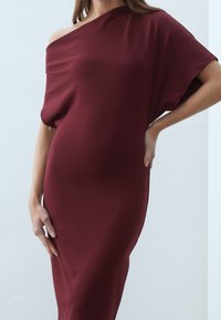 Robe bordeaux, dénudée d'épaule, en tissu doux, avec une coupe décontractée et un design drapé, reposant juste au-dessus de la cheville.