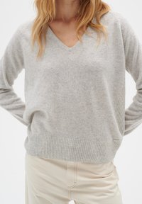 Femme portant un pull gris clair à col en V avec ourlet et poignets côtelés, associé à un pantalon beige clair, visible du cou jusqu'aux hanches.