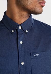 Man som bär en marinblå skjorta med knappar i vitt och en ficka med en liten broderad fågel-logotyp.