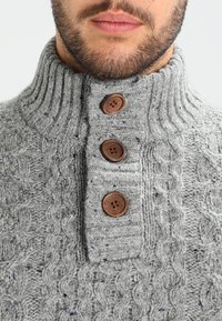 Suéter gris de punto con un alto cuello acanalado, que presenta tres botones marrones y un patrón de cable texturizado en toda su superficie.