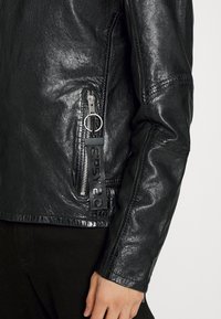 Czarna skórzana kurtka z rękawem i kieszenią boczną na zamek błyskawiczny z metalowym uchwytem oraz metką "GIPSY 2.0", noszona na czarnych spodniach.