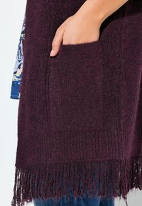 Hand teilweise in der Tasche eines strukturierten, dunkellila gestrickten Schals mit Fransen, über einem blauen gemusterten Ärmel und Jeans.