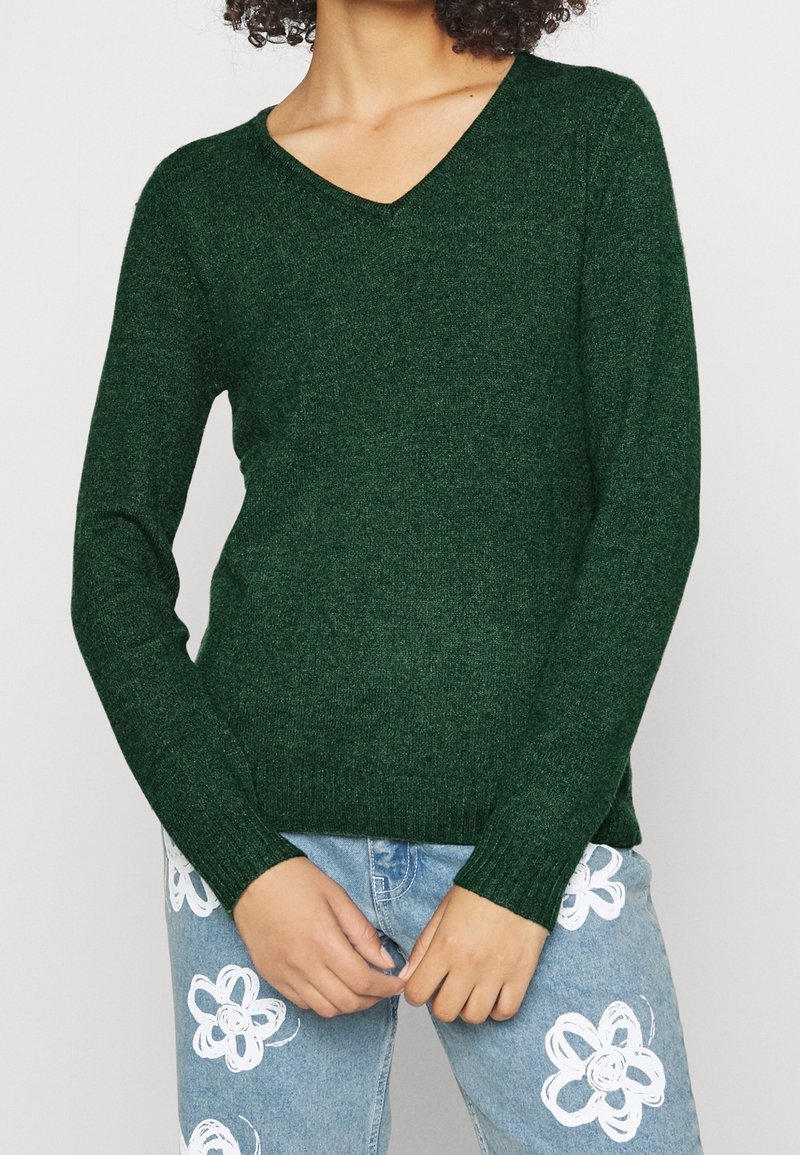 Grüner Strickpullover mit V-Ausschnitt und langen Ärmeln, der eine strukturierte Oberfläche aufweist. Kombiniert mit hellblauen Jeans, die mit weißen Blumenmustern verziert sind.