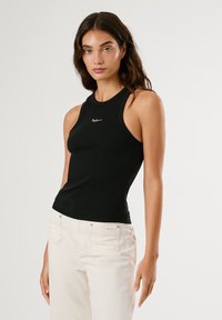 Pikkade pruunide juustega naine, kes kannab musta varrukateta topi ja valgeid pükse, seisab ühevärvilise heleda tausta ees.