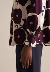 Blusa a maniche lunghe color crema con grande motivo floreale bordeaux. Presenta polsini arricciati con dettaglio a bottone. Tessuto liscio con una consistenza morbida.