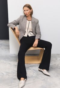 Grauer karierten Blazer über einem beigen Oberteil mit schwarzen Akzenten, kombiniert mit schwarzen ausgestellten Hosen und hellfarbigen Slippern. Minimalistischer Holzstuhl.