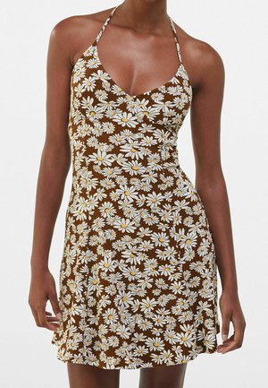 Vestido halter marrón sin mangas con estampado de margaritas blancas y amarillas, llevado por una persona con los brazos relajados a los lados, sobre un fondo claro liso.