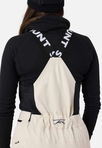 Beige og sorte skidragter med krydsryg-design, der har elastiske stropper med "SWEDEMOUNT" og synlig tekstureret stof.