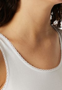 Witte tanktop met een geribbelde textuur en een delicate kantafwerking langs de halslijn, die een gladde, aansluitende pasvorm accentueert.