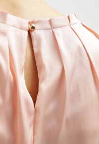 Vaaleanpunainen pusero, jossa on poimuttavaa kangasta, avaus niskassa, jossa on avainreikä, ja pyöreä kultasävyinen nappisulkija. Silkkinen rakenne.