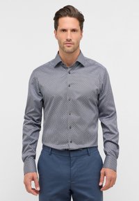 Overhemd met knopen in een patroon van stof, met een blauwe basis en kleine geometrische ontwerpen, lange mouwen en bruine knopen. De broek is marineblauw.