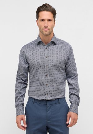 Camicia button-up in tessuto stampato, con una base blu e piccoli disegni geometrici, maniche lunghe e bottoni marroni. I pantaloni sono di colore blu navy.
