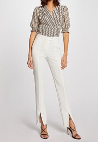 Top envolvente negro y beige con estampado y mangas abullonadas, combinado con pantalones blancos acampanados con aberturas laterales y zapatos de tacón negro con tiras.