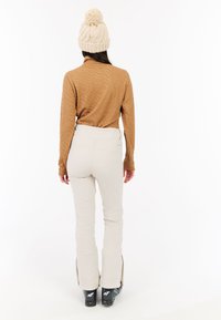 Smeđi strukturirani dukserica dugih rukava u kombinaciji s off-white skijaškim hlačama koje imaju patentne zatvarače na gležnjevima, nosi se s crnim skijaškim čizmama i bež pletenom kapom.