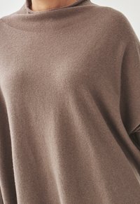 Dziany poncho w ciepłym kolorze taupe o miękkiej fakturze, z wysokim dekoltem i luźnym krojem, zaprojektowane z myślą o komforcie i swobodzie ruchów.