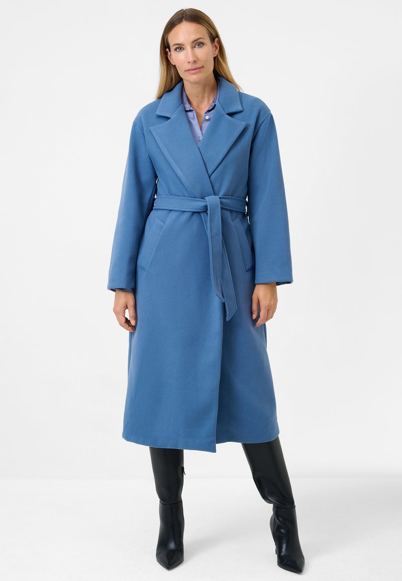 Cappotto blu con cintura realizzato in tessuto testurizzato, caratterizzato da un colletto a risvolto, maniche ampie e due tasche frontali, abbinato a stivali aderenti neri.