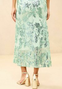 Mint green skirt with intricate sequin floral patterns, flowing design, sheer layering, paired with gold block-heeled sandals.