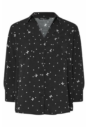 Zwarte blouse met witte stippen en kleine strikvormen, voorzien van een kraag en twee voorzakken. De stof lijkt zacht en lichtgewicht.