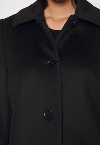 Abrigo negro con un cuello estructurado, que presenta dos grandes botones negros y un acabado texturizado. Diseño simple y clásico.