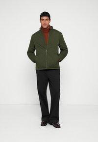 Green quilted jacket with a high collar and front zipper. Worn over a rust-colored turtleneck, paired with black trousers and shoes.