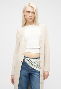 Ξανθιά γυναίκα που φοράει ένα μακρύ μπεζ ζακέτα, λευκό κοντό τοπ και μπλε τζιν με αξεσουάρ λευκή πλεκτή ζώνη.