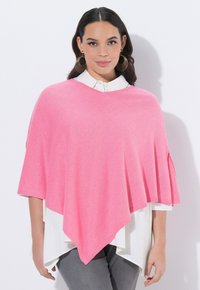 Růžový pončový svetr z měkké látky s výstřihem do V a krátkými rukávy, vrstvený přes bílou košili s límečkem a šedé kalhoty.