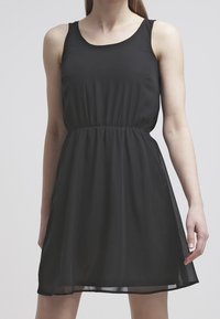 Musta hihaton mekko, jossa on pyöreä pääntie, joustava vyötärö ja virtaava, läpikuultava kangas, joka ulottuu polvien ylle.