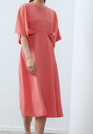Vrouw draagt een koraalroze midi-jurk met losse korte mouwen en staat in een minimalistische witte binnenruimte.