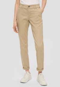 Femme portant un pantalon chino beige retroussé et des baskets blanches, debout avec les mains détendues le long du corps, sur un fond uni.