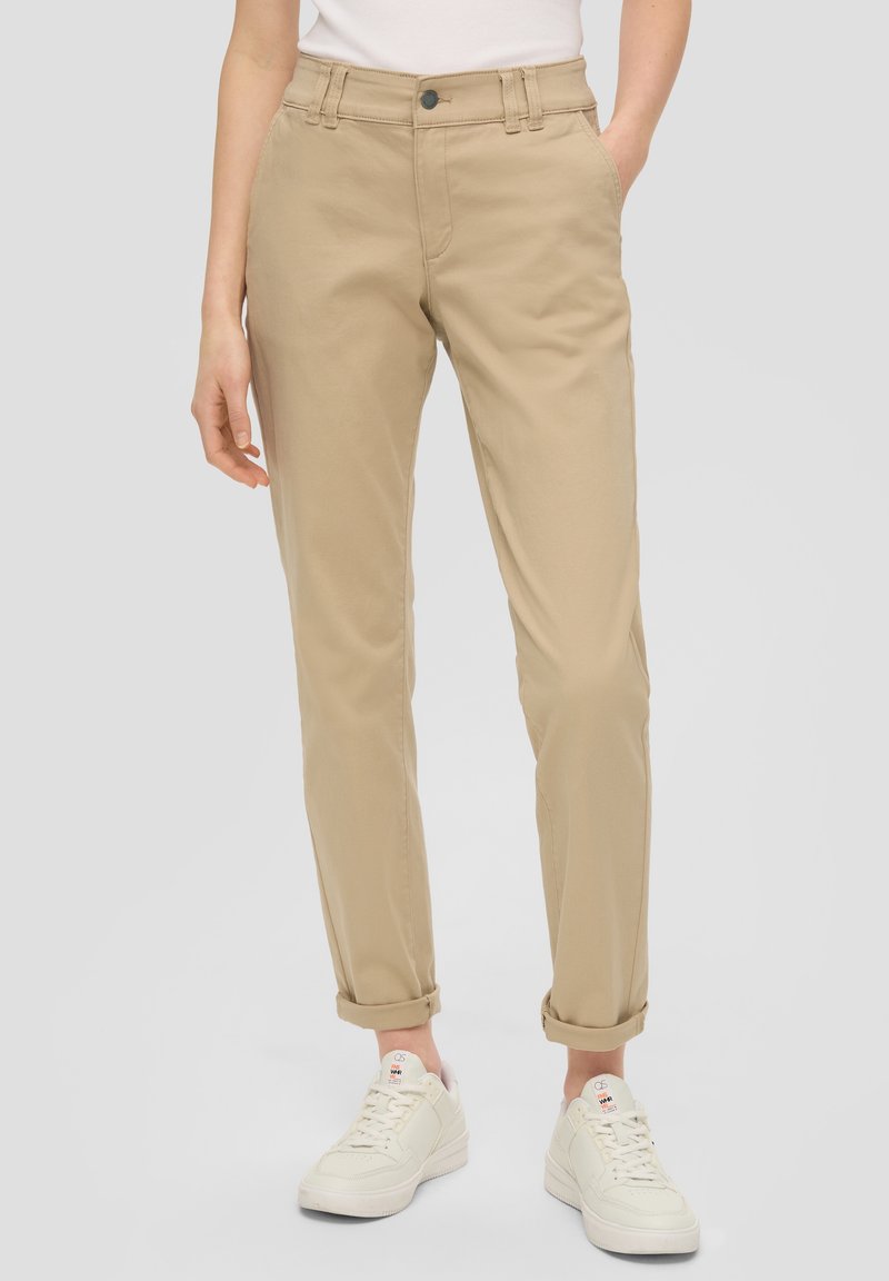 Femme portant un pantalon chino beige retroussé et des baskets blanches, debout avec les mains détendues le long du corps, sur un fond uni.