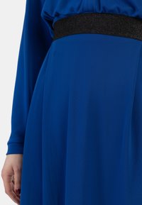Blå kjole med en flydende silhuet, lange ærmer og en tekstureret sort talje. Stoffet har et glat, blødt udseende.