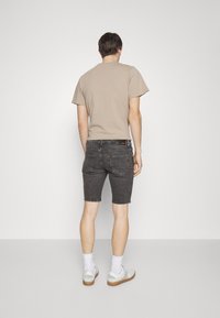 En ung man står med ryggen mot kameran, iklädd en beige t-shirt, mörka shorts och vita sneakers, framför en enfärgad bakgrund.