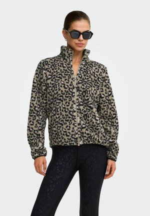 Žena nosiaca fleecovú bundu na zips s leopardím vzorom a čierne legíny s jemným zvieracím vzorom, stojaca pred jednoduchým svetlým pozadím.