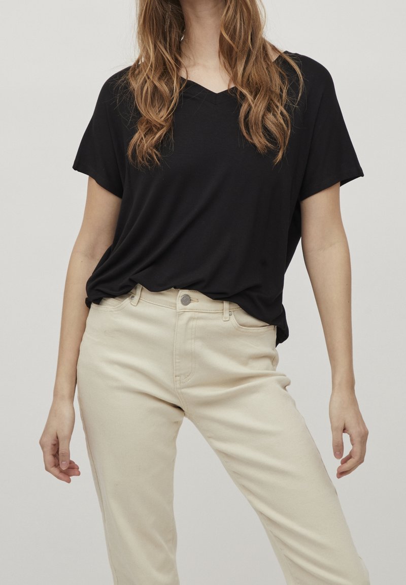 Kvinna som bär en lös svart kortärmad V-ringad topp instoppad i högmidjade beiga byxor mot en enfärgad bakgrund.