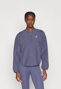 Giacca leggera viola con zip frontale, polsini elasticizzati e texture verticale a righe. Presenta un logo sul petto sinistro.