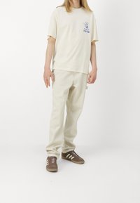 Camisa de algodón beige con un gráfico de un gato azul en el pecho, combinada con pantalones beige claros y zapatillas marrones con rayas blancas.