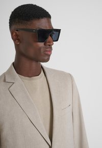 Occhiali da sole neri oversize con montatura geometrica e lenti oscurate, abbinati a un blazer di lino beige sopra un top in maglia coordinato.