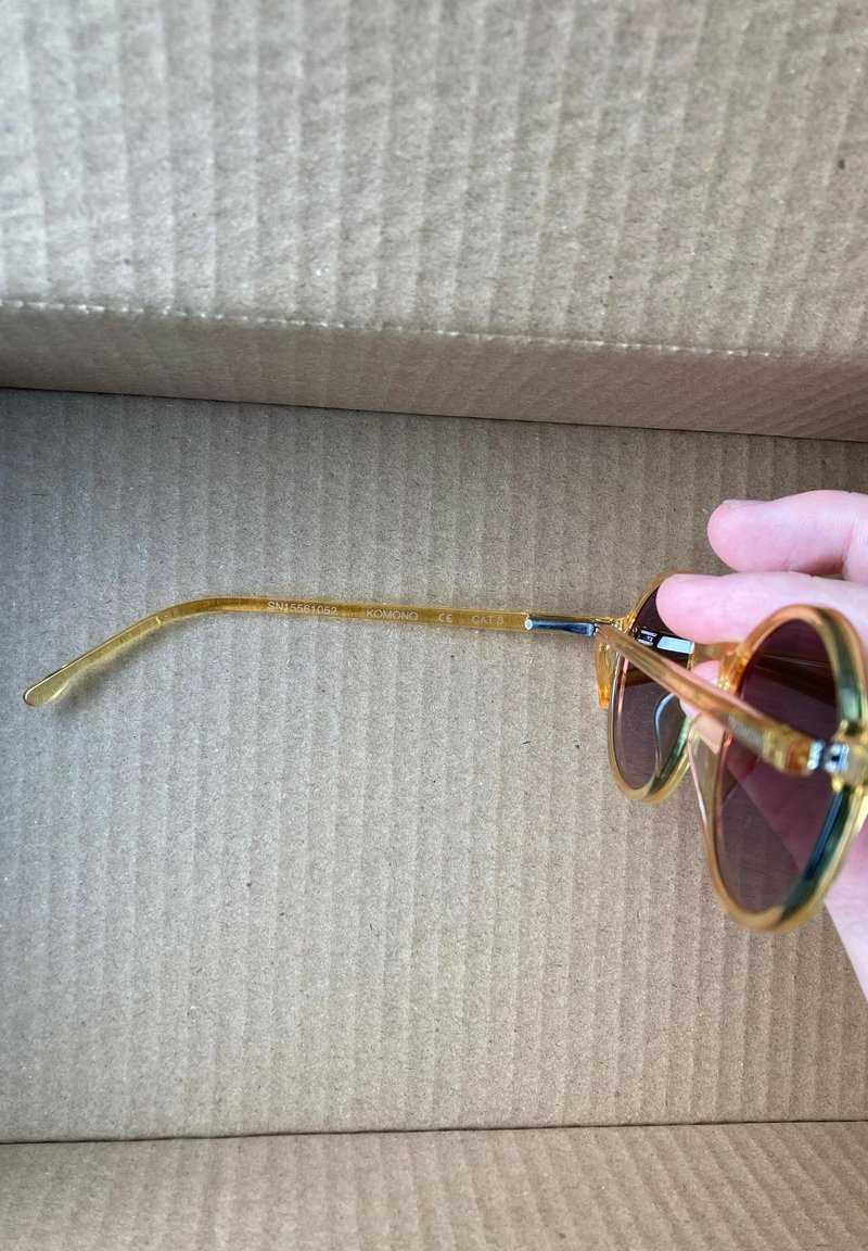 Lunettes de soleil rondes avec des montures transparentes orange, des verres teintés foncés et des branches flexibles, reposant sur une surface en carton texturé.