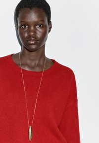 Czerwony dzianinowy sweter z okrągłym dekoltem, noszony na złotym naszyjniku z wisiorkiem, o gładkim, wydłużonym kształcie. Prosta, teksturowana tkanina.